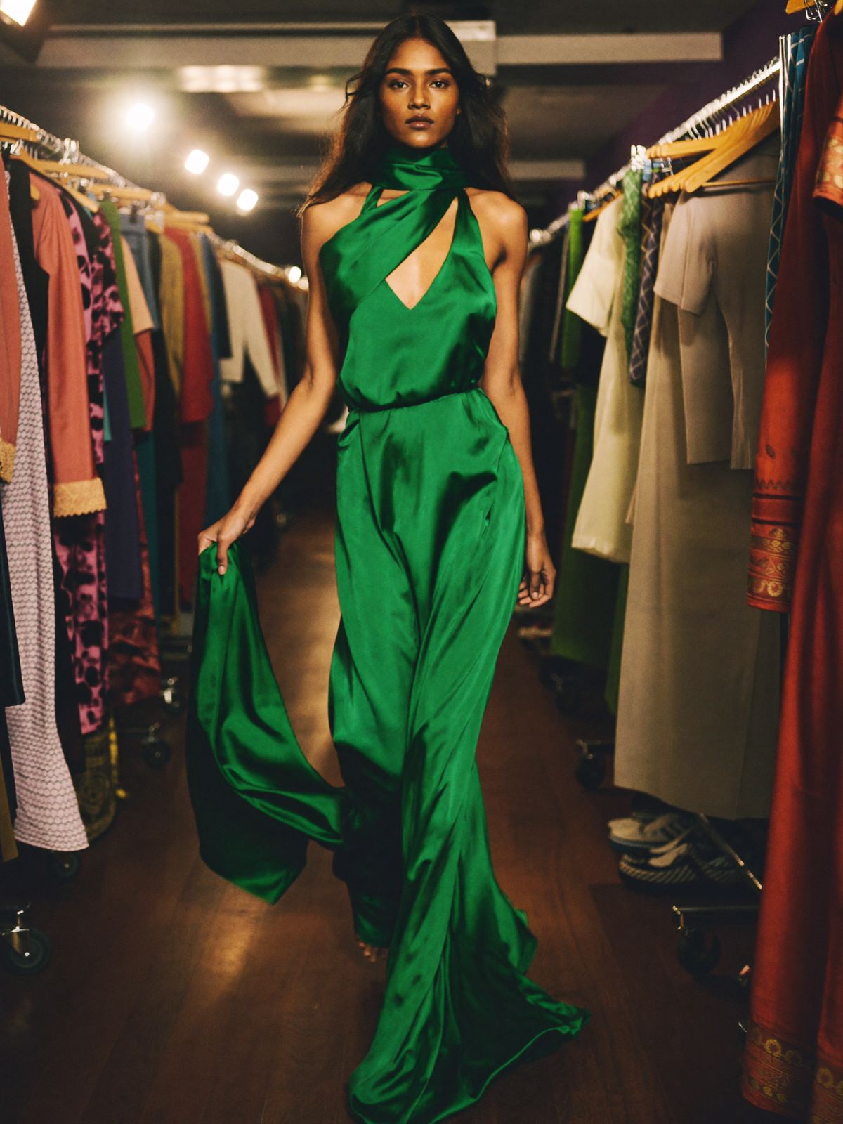 A model in an emerald satin gown walking backstage between rails of clothes